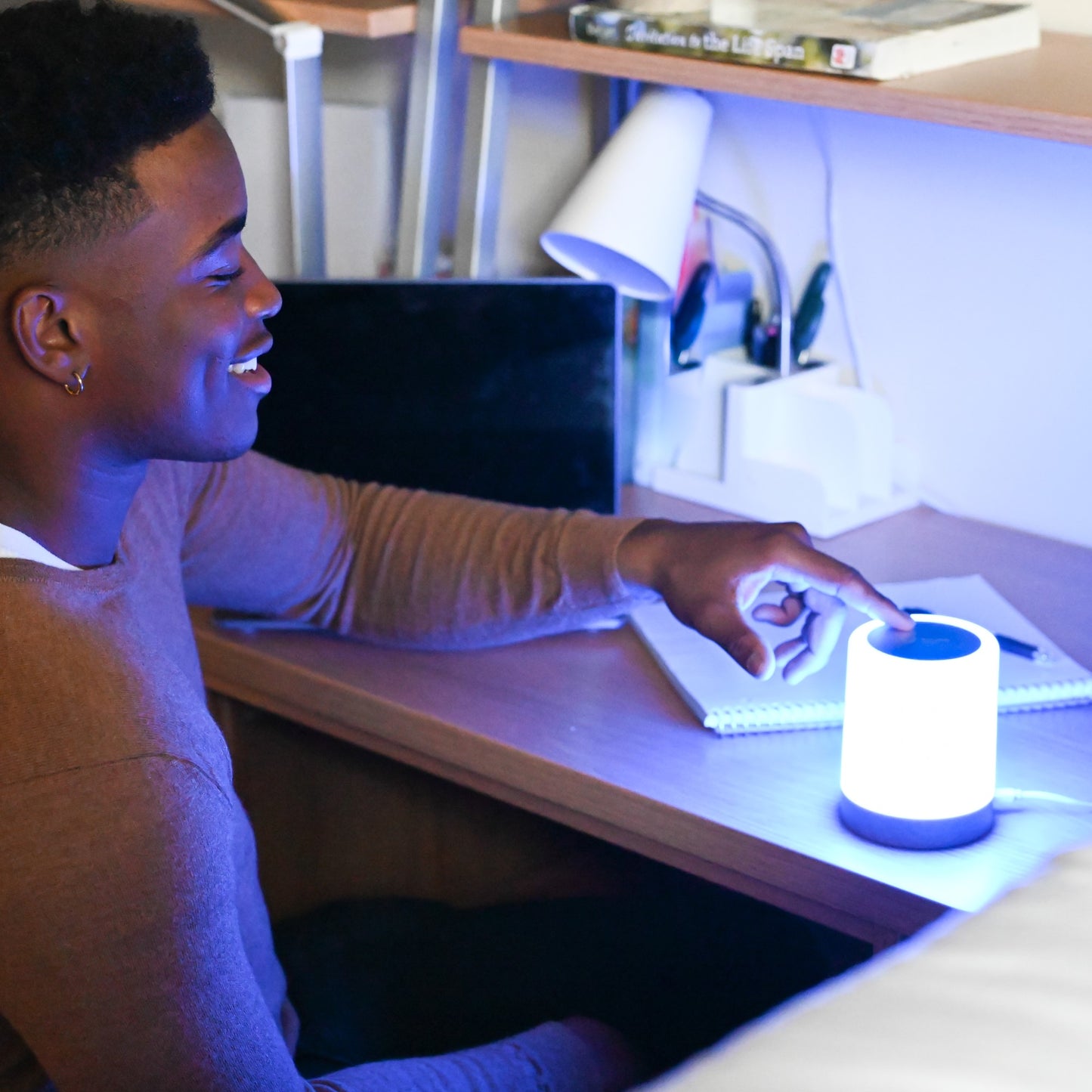 Person smiling while touching a glowing white and blue LuvLink Friendship Lamp on their desk at night.