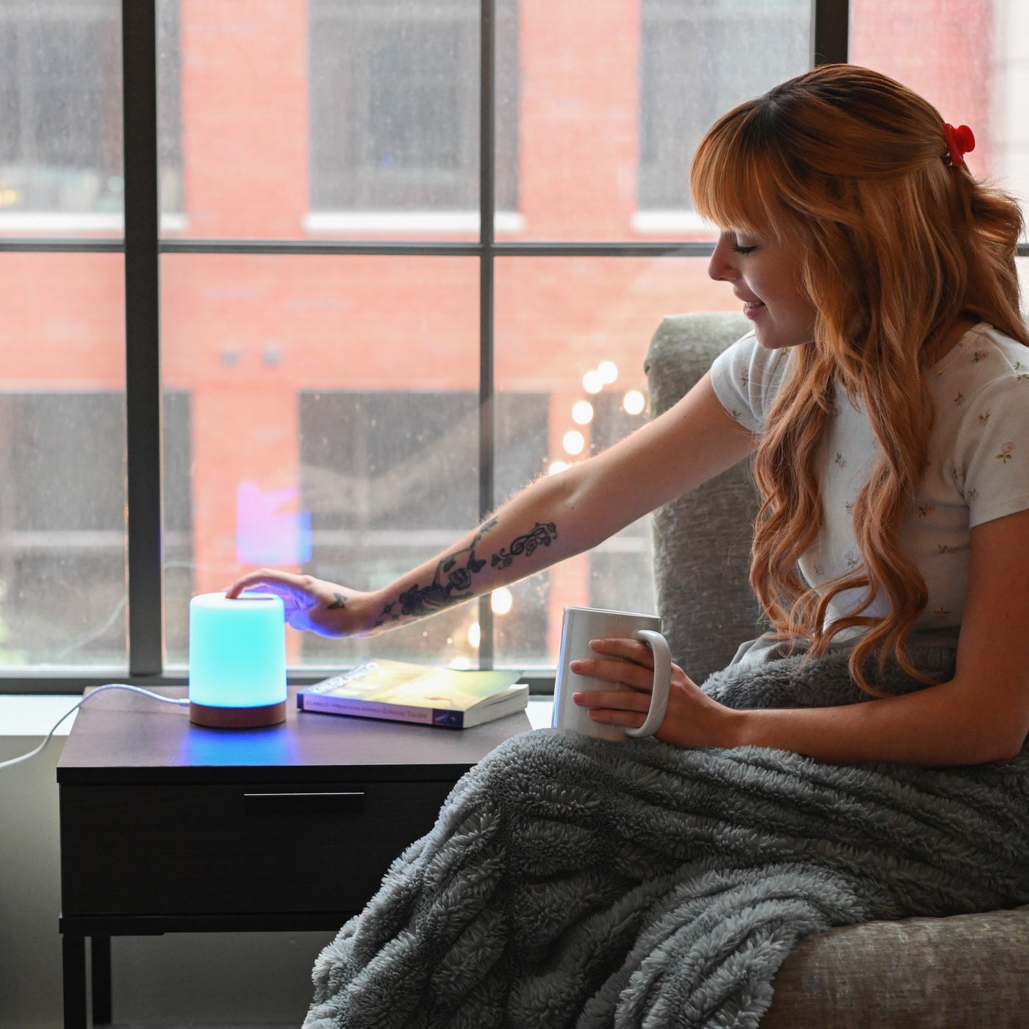 Woman touching a glowing blue LuvLink Friendship Lamp while holding a mug, sitting by a window overlooking a city.