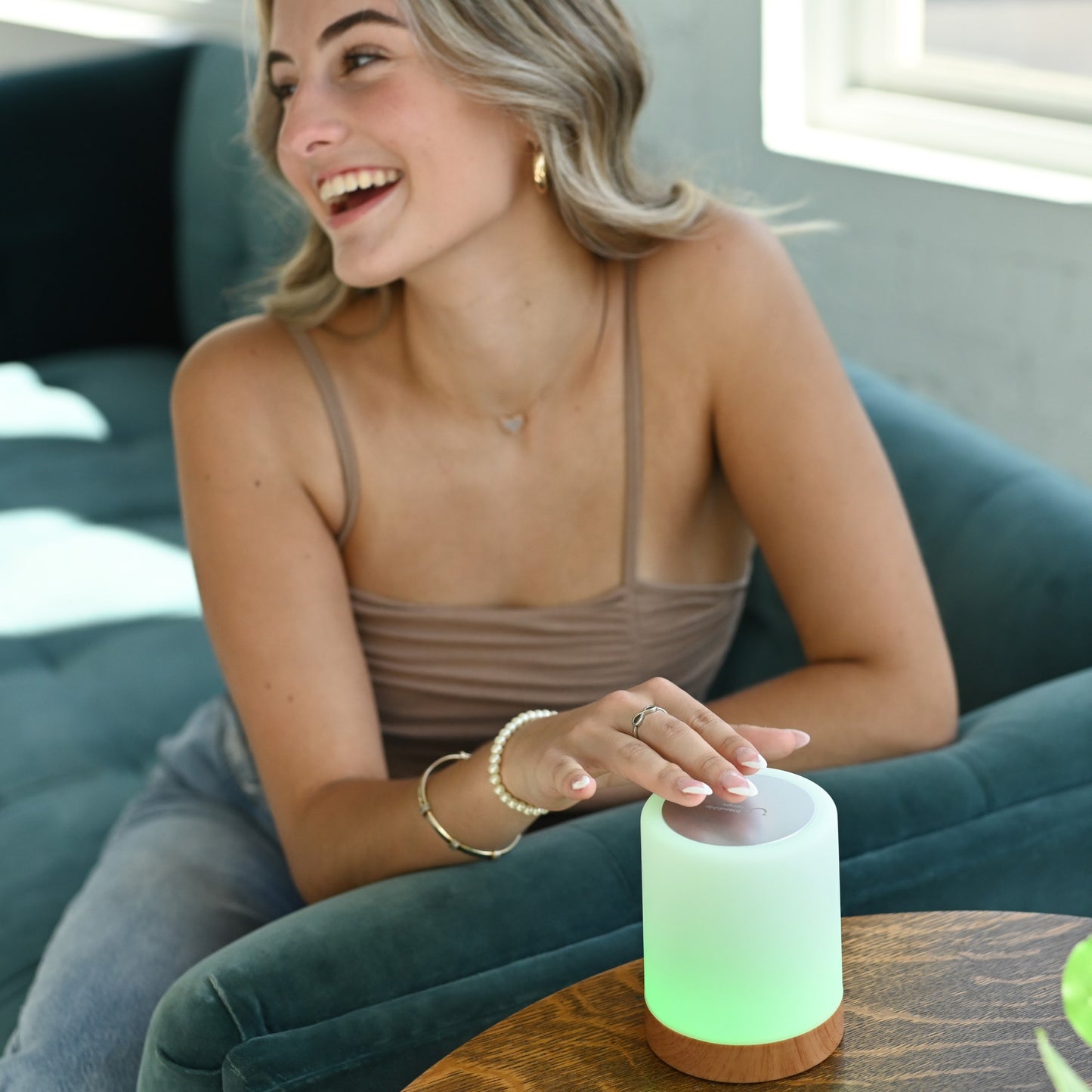 Woman smiling while touching a glowing green LuvLink Friendship Lamp on a wooden table in a modern living room.