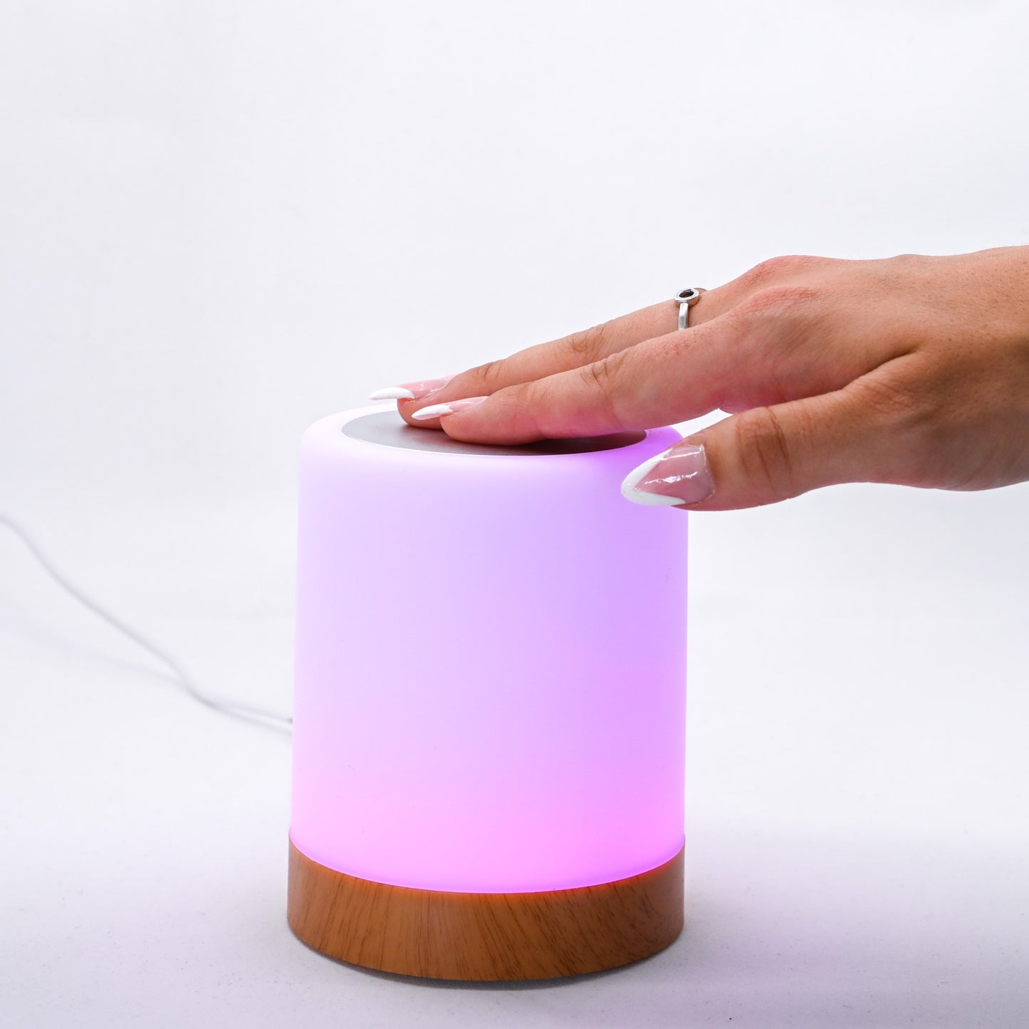 Hand touching the top of a glowing pink LuvLink Friendship Lamp with wooden base against white background.